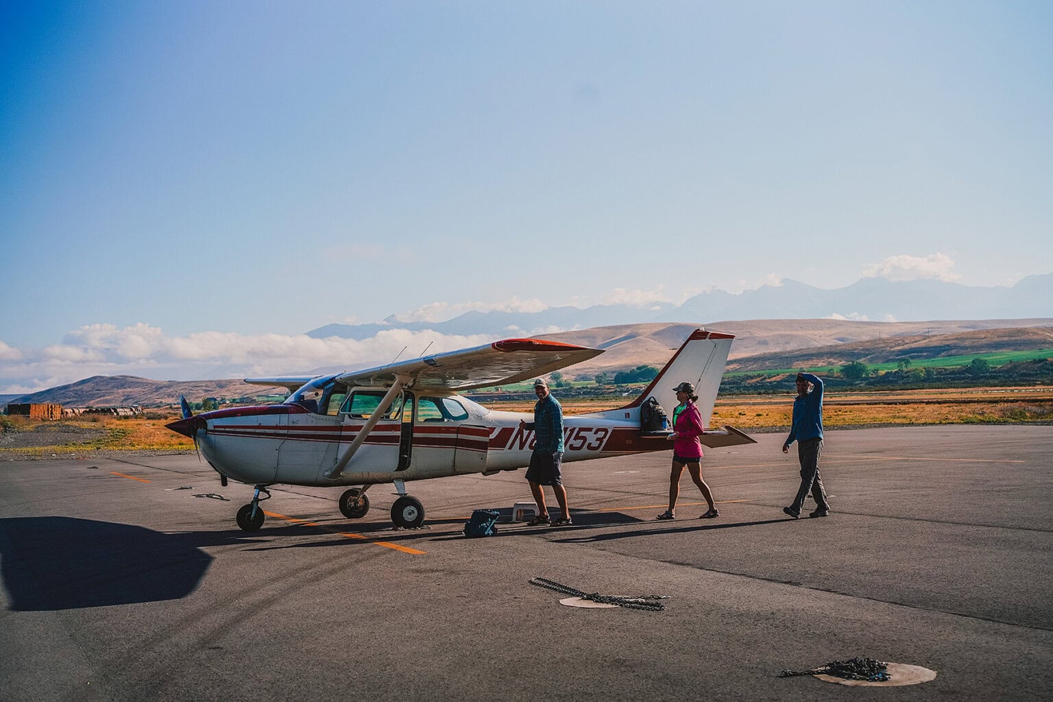 Three people approach a small plane on a scenic airstrip in Idaho with rolling hills in the distance.