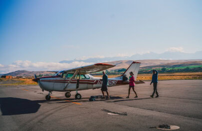 Three people approach a small plane on a scenic airstrip in Idaho with rolling hills in the distance.