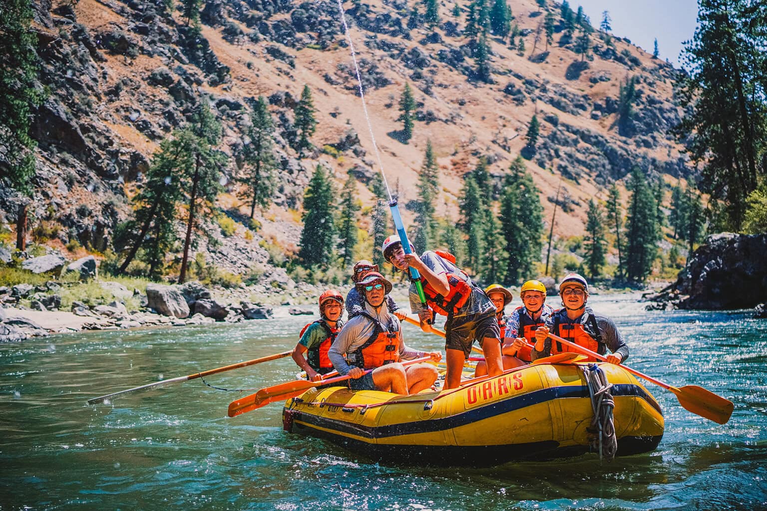 A group of people in life jackets paddles a yellow raft down a river surrounded by rocky hills and tall evergreen trees. A boy in front sprays a water gun in the air.