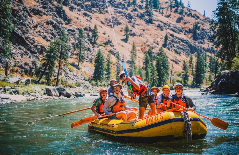 A group of people in life jackets paddles a yellow raft down a river surrounded by rocky hills and tall evergreen trees. A boy in front sprays a water gun in the air.