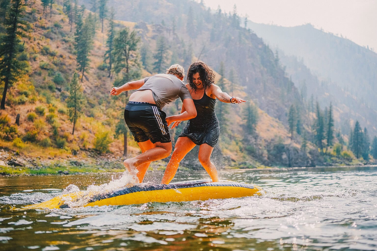 Two people playing on top of an upside down inflatable kayak in the middle of a river, trying to knock each other off
