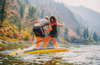 Two people playing on top of an upside down inflatable kayak in the middle of a river, trying to knock each other off