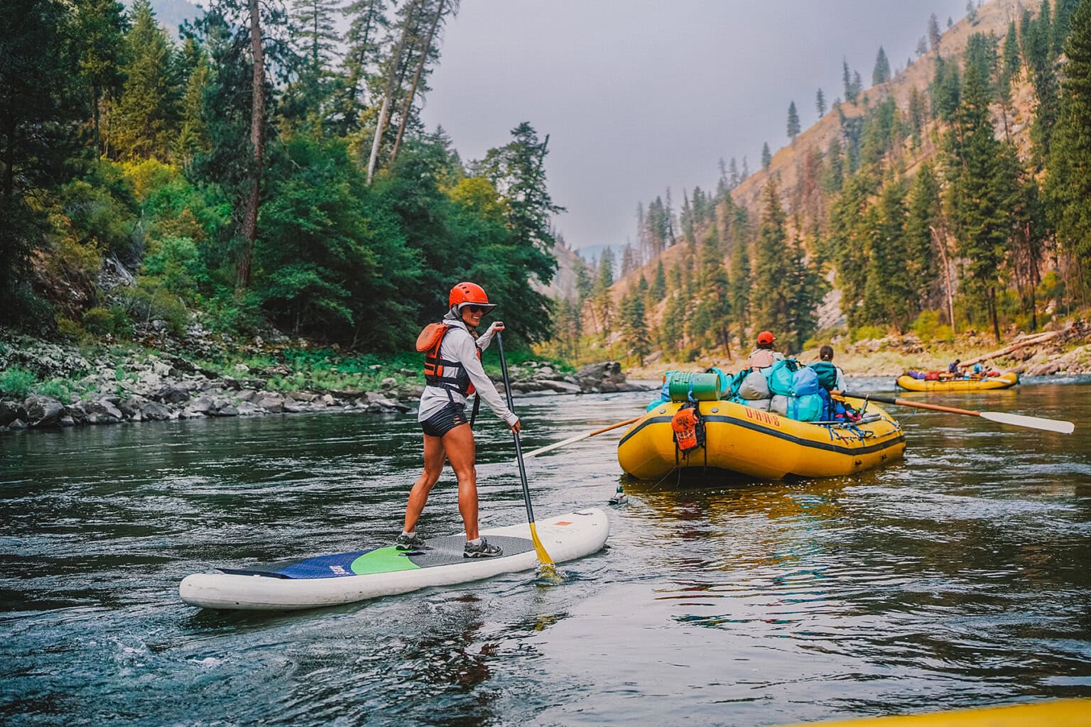 A woman paddles a stand up paddleboard behind a raft on a scenic stretch of the Main Salmon River surrounded by forest and mountainous terrain