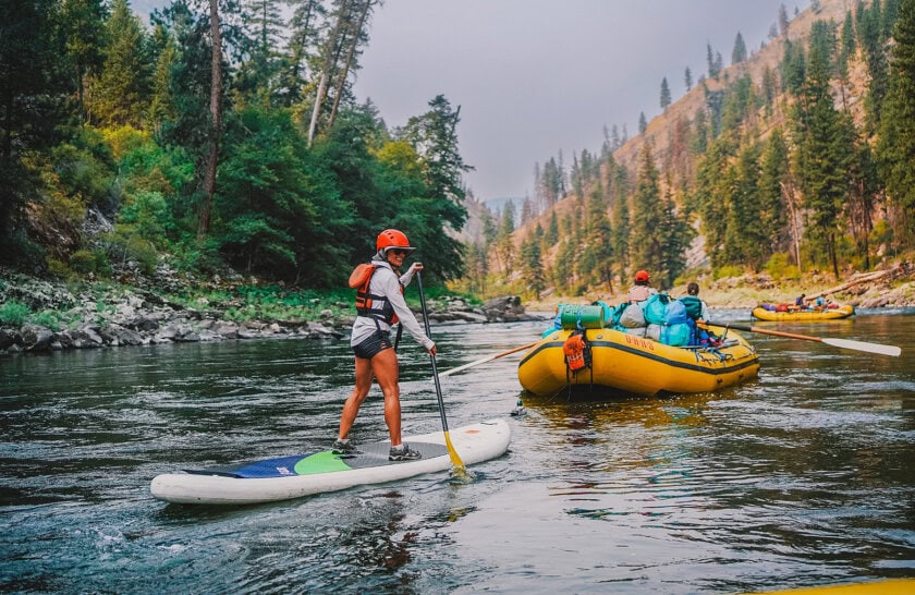 A woman paddles a stand up paddleboard behind a raft on a scenic stretch of the Main Salmon River surrounded by forest and mountainous terrain