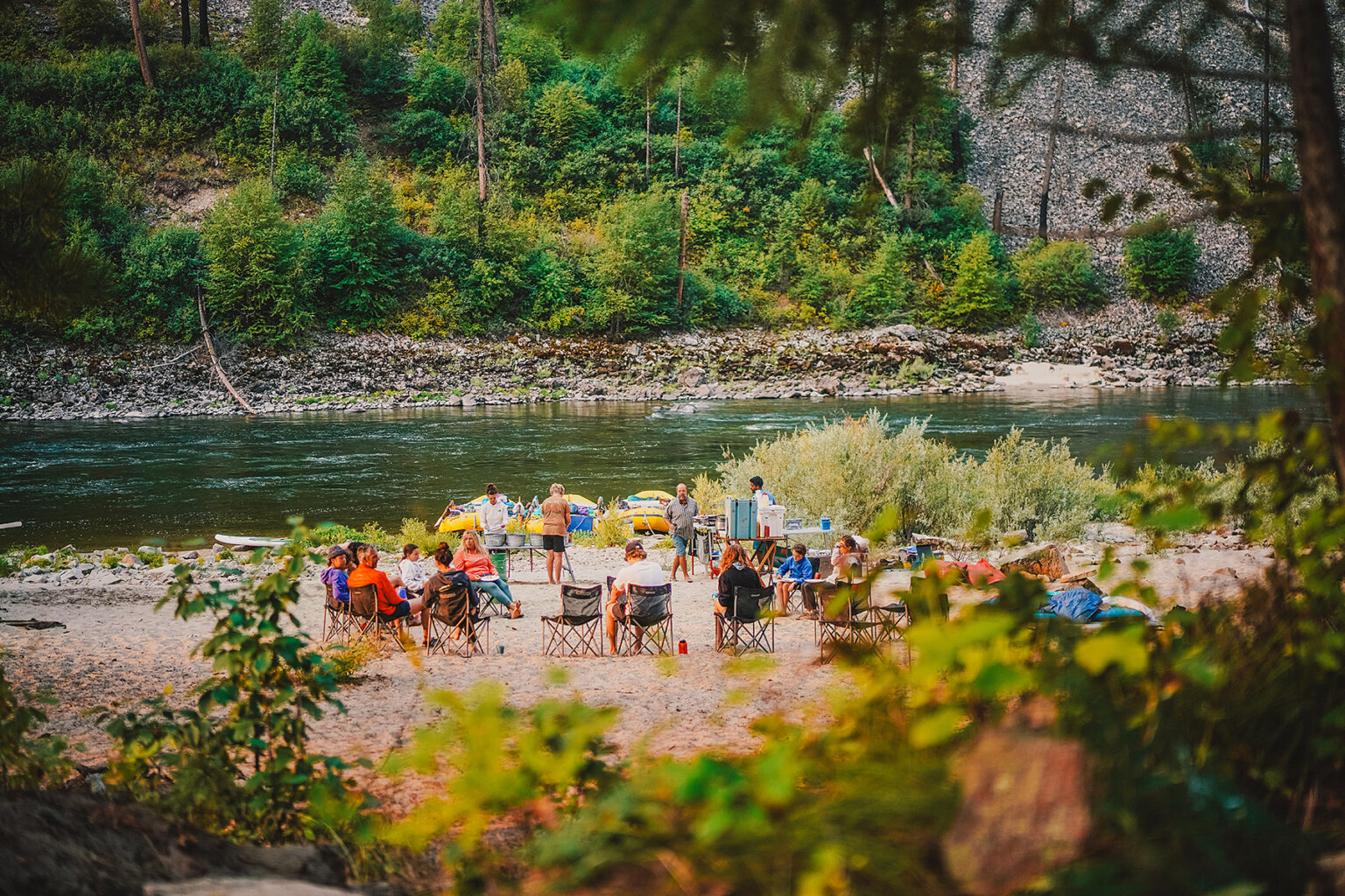 A group of rafters enjoying some downtime sits in chairs sprawled around a beach camp setting alongside a scenic river