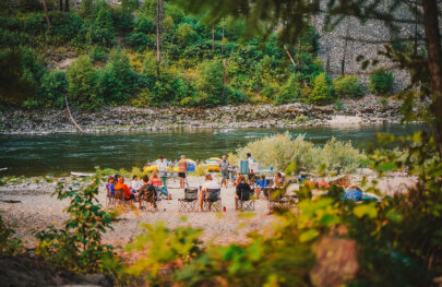 A group of rafters enjoying some downtime sits in chairs sprawled around a beach camp setting alongside a scenic river