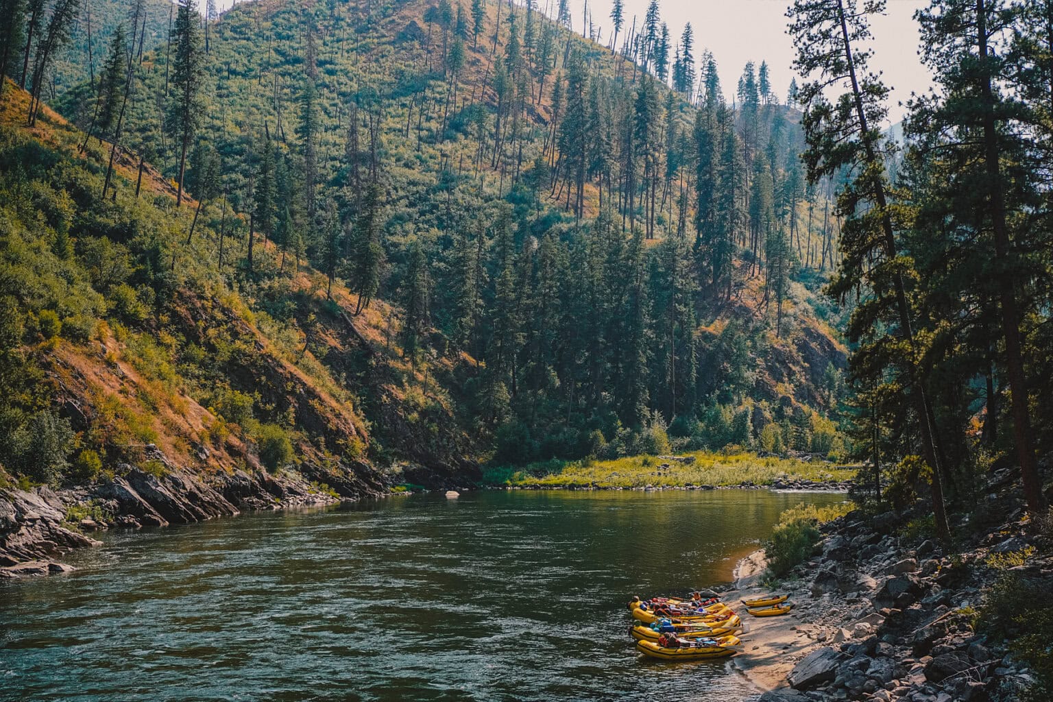 A serene river scene with several yellow rafts on a sandy bank, surrounded by lush green hills and tall trees under a hazy sky.