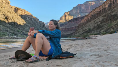 A woman sits on a sandy beach next to the Colorado River with towering cliffs rising behind her in Grand Canyon