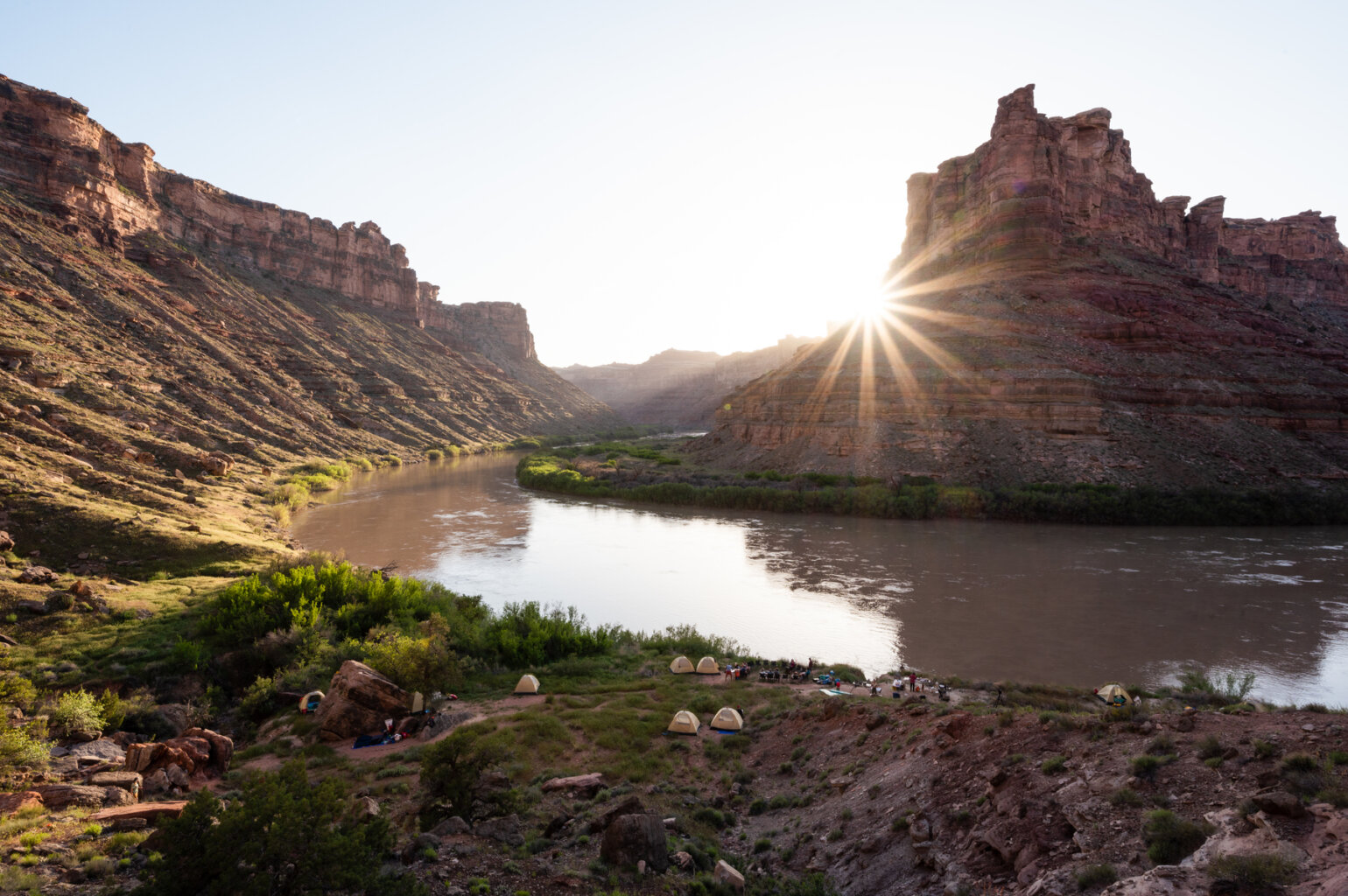 Camp set up along the Colorado River in Cataract Canyon as the sun sets behind canyon walls creating a sunburst.