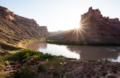 Camp set up along the Colorado River in Cataract Canyon as the sun sets behind canyon walls creating a sunburst.