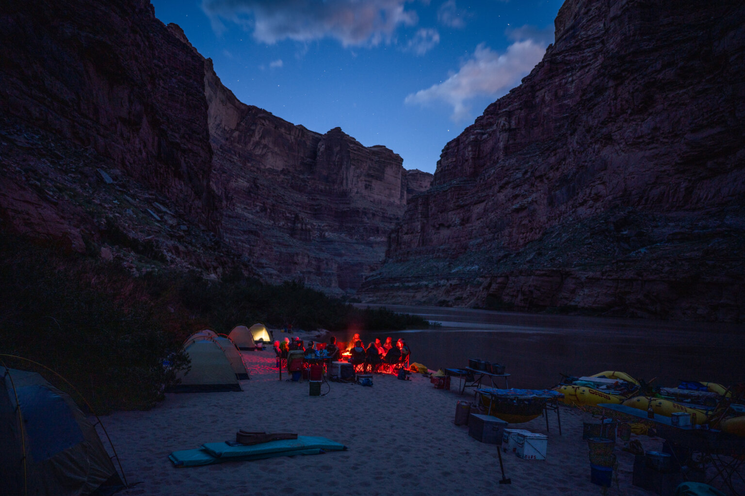 Several guests sit around a glowing red campfire at camp during twilight along the Colorado River through Cataract Canyon.