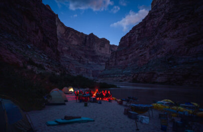 Several guests sit around a glowing red campfire at camp during twilight along the Colorado River through Cataract Canyon.