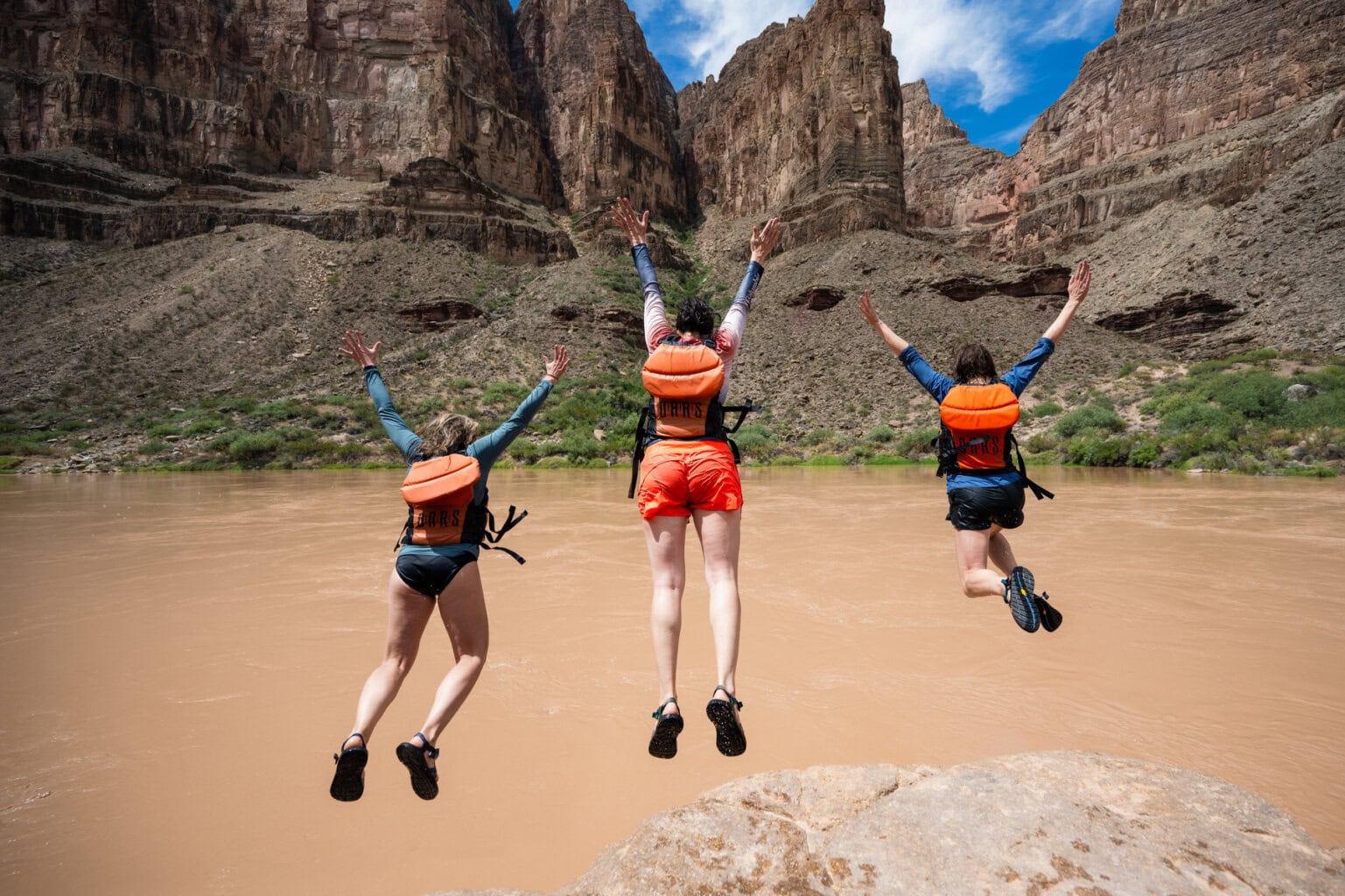 Three guests jump off a rock with their hands in the air into the Colorado River in Grand Canyon