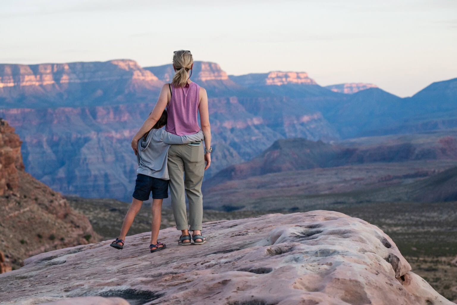 A mom and daughter hold on to each other as they look out over a vast canyon on the rim of Grand Canyon