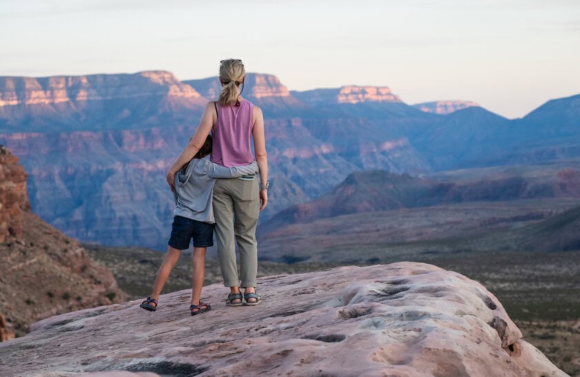 A mom and daughter hold on to each other as they look out over a vast canyon on the rim of Grand Canyon