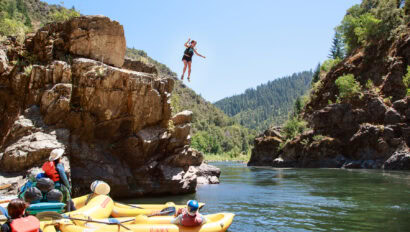 A woman jumps from a cliff into a river where rafts wait