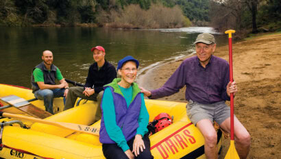 A family of four sits on a yellow raft on shore next to a river in a forested setting
