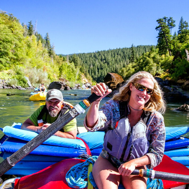 A smiling guide rows downstream on the Rogue River with a happy guest leaning on the pile of sleeping pads stacked up behind her.