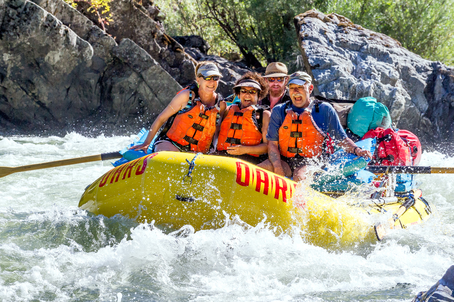Three people sit in the front of a yellow raft as a guide rows it through a rapid with ecstatic smiles.