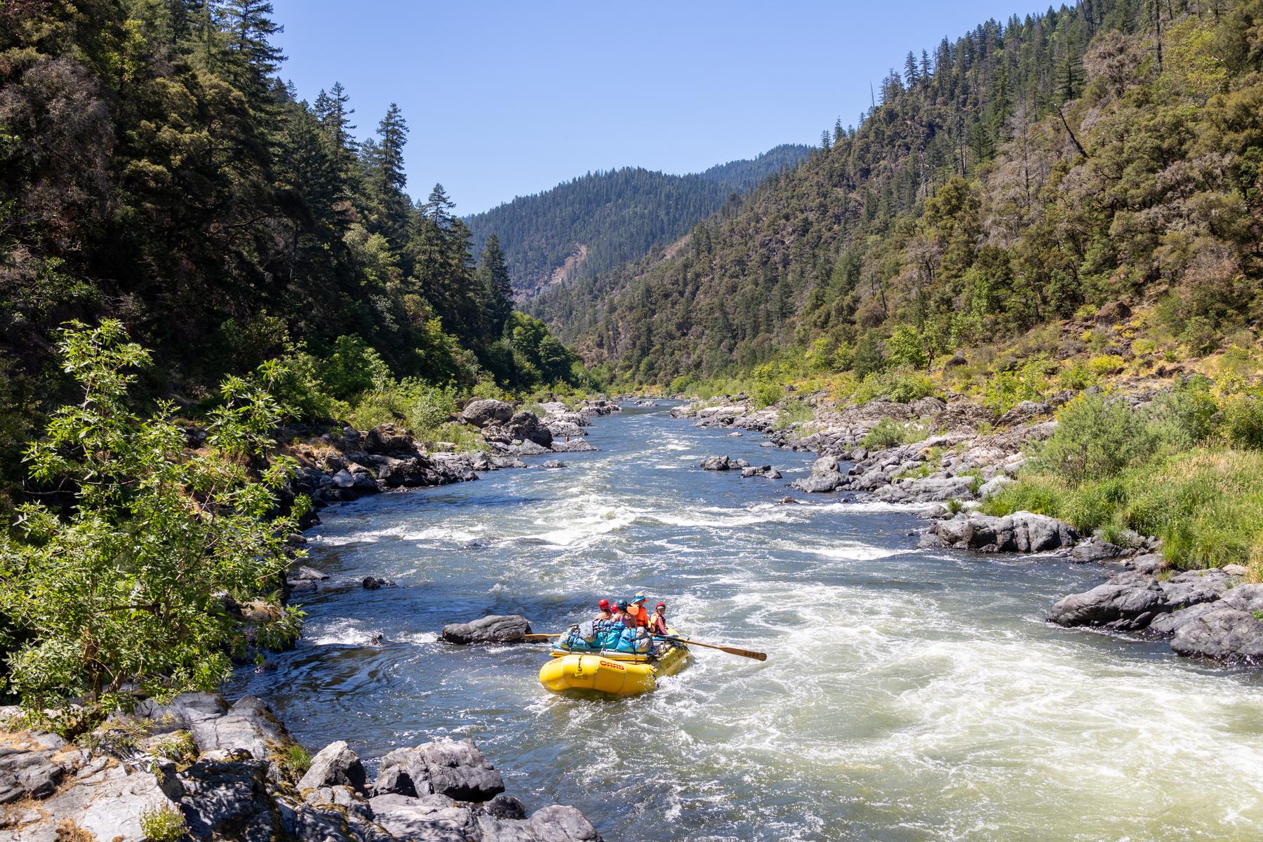 A yellow raft with four people navigates the Wild & Scenic Rogue River surrounded by steep green hills and rocky shores under a clear blue sky.