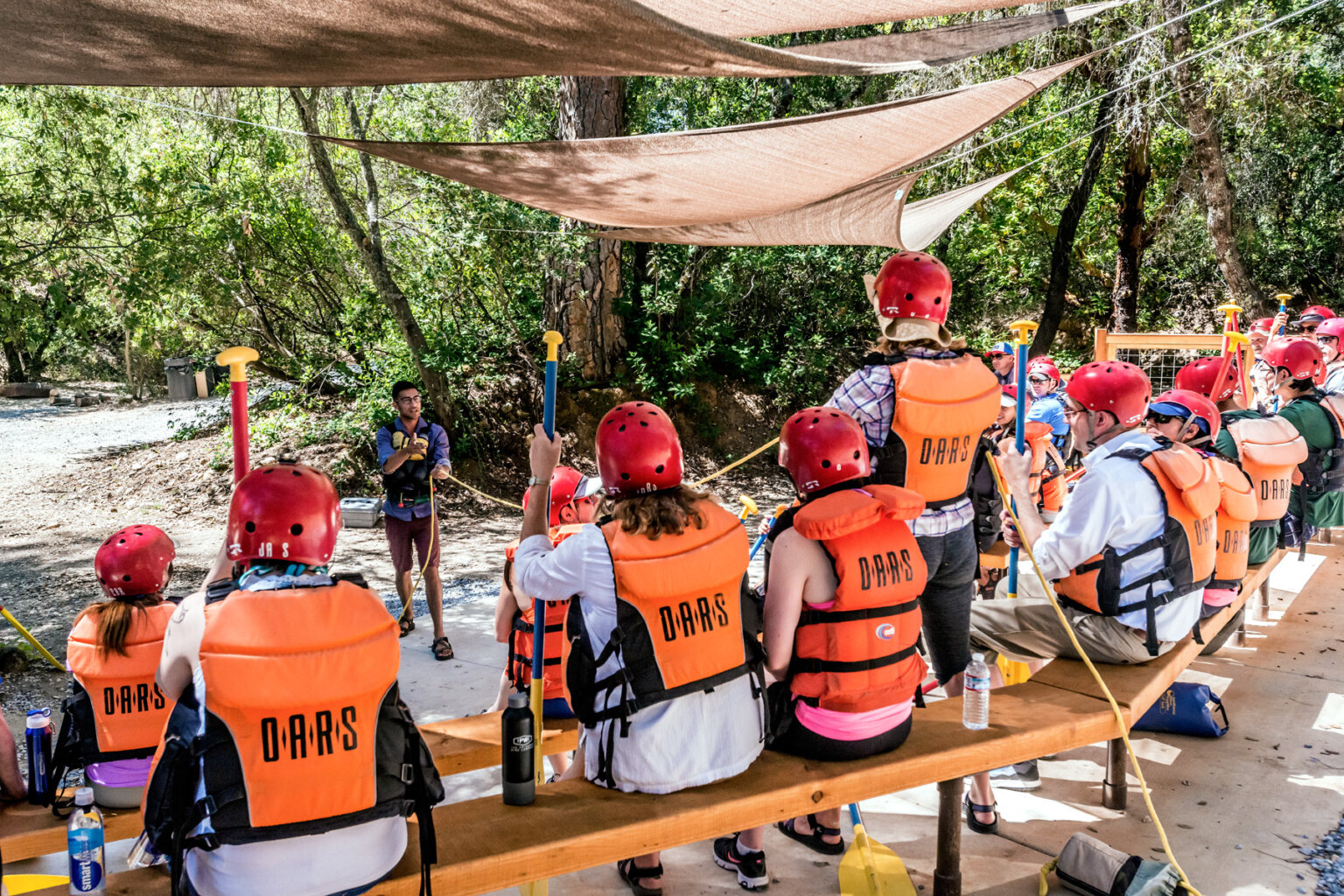 A guide conducts a safety talk as guests in personal floatation devices and helmets sit and watch at the OARS American River Outpost.