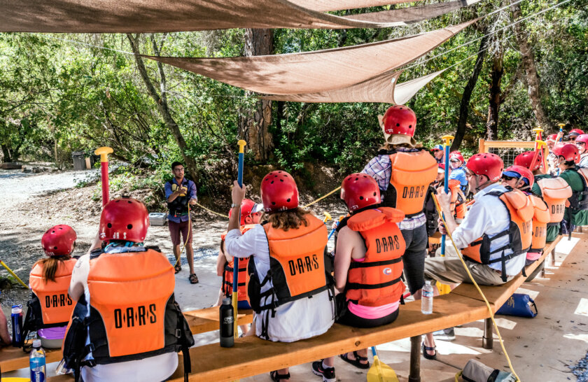 A guide conducts a safety talk as guests in personal floatation devices and helmets sit and watch at the OARS American River Outpost.