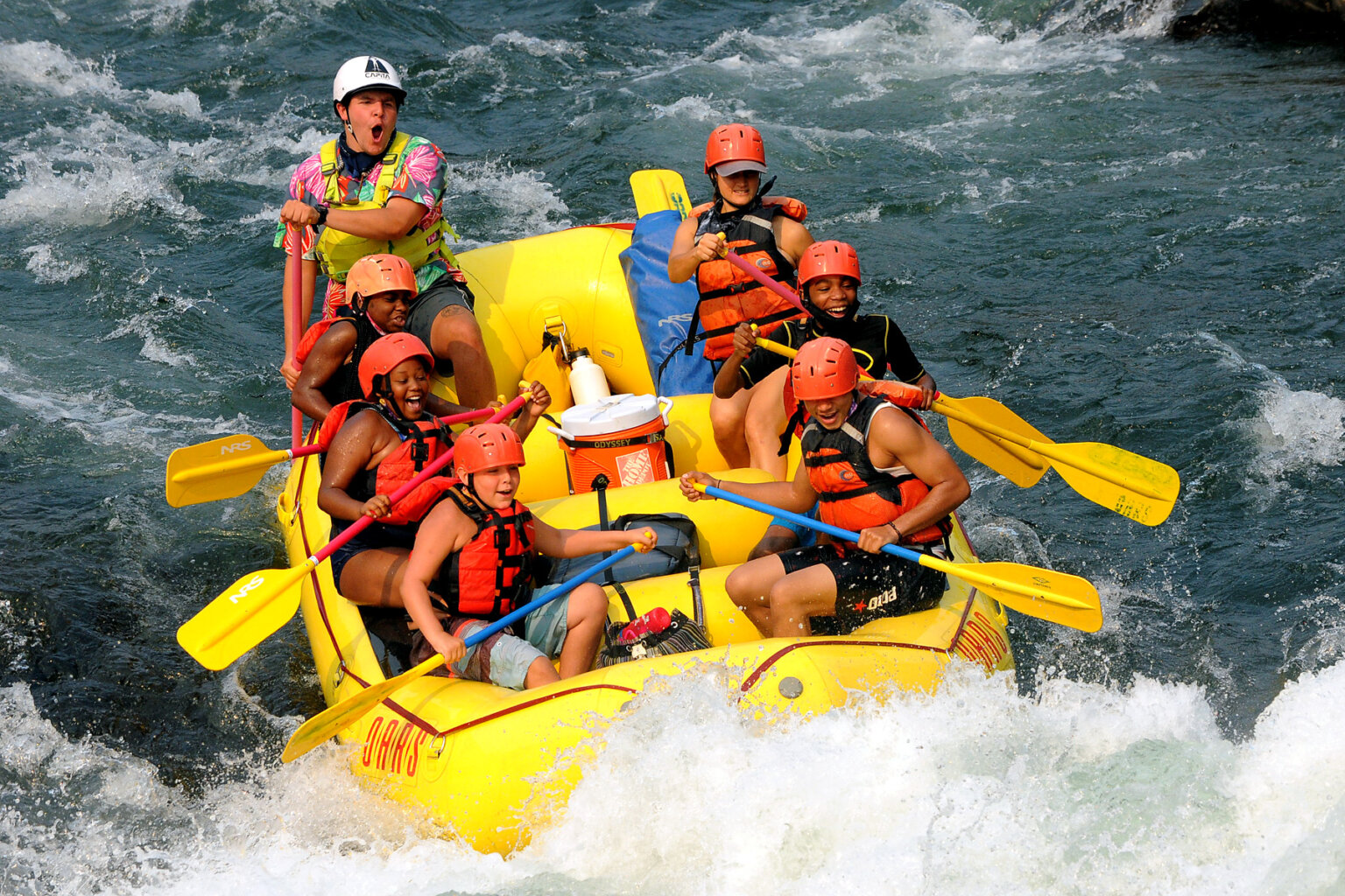 Six excited young guests hold on to their paddles as their guide paddles them through a rapid.