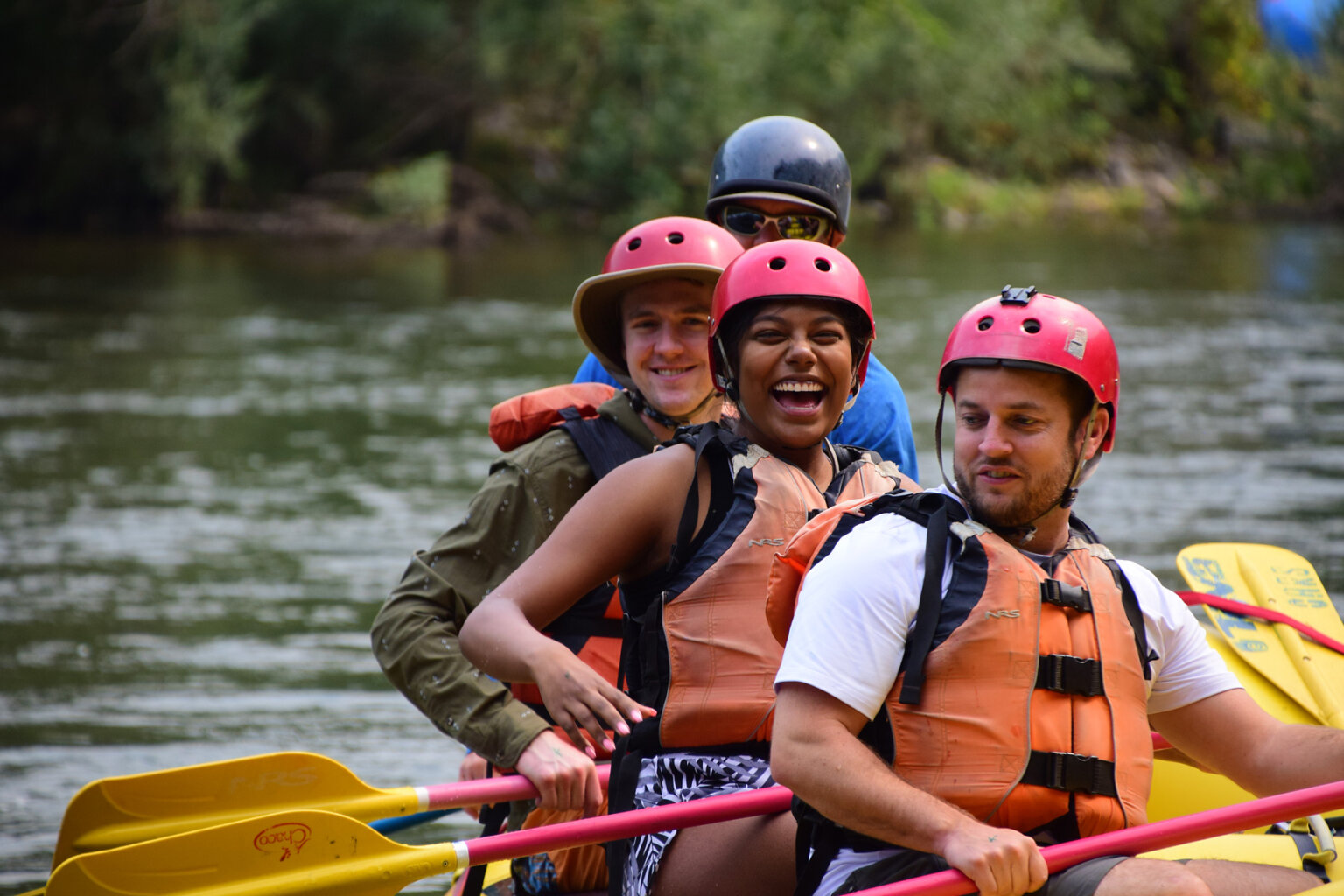 A woman smiles ecstatically at the camera in a line of three guests as they sit on a yellow raft with paddles ready to row.