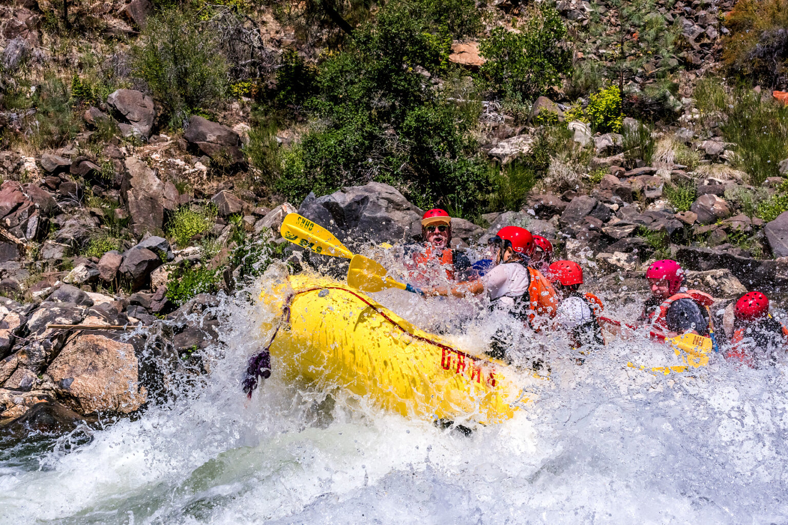 A yellow raft crashes through a rapid and the paddlers are getting soaked by wave spray.