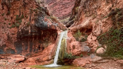 A waterfall spills over a ledge from a vibrant red rock canyon