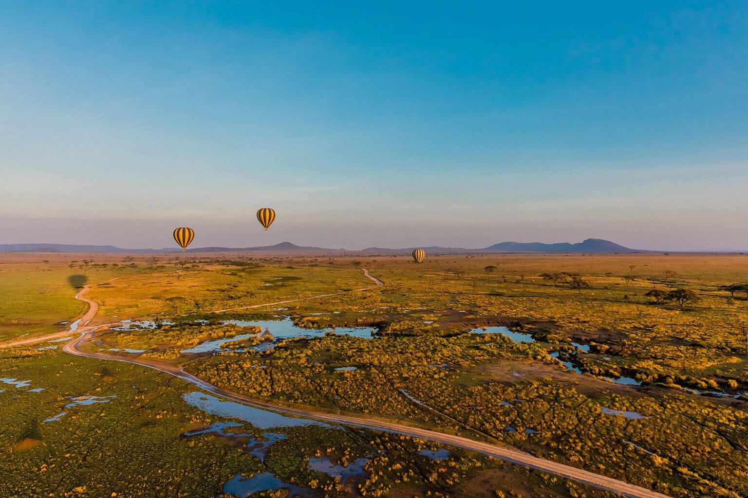 Aerial view of a vast landscape with three striped hot air balloons drifting over greenery and a winding dirt road, under a clear blue sky.