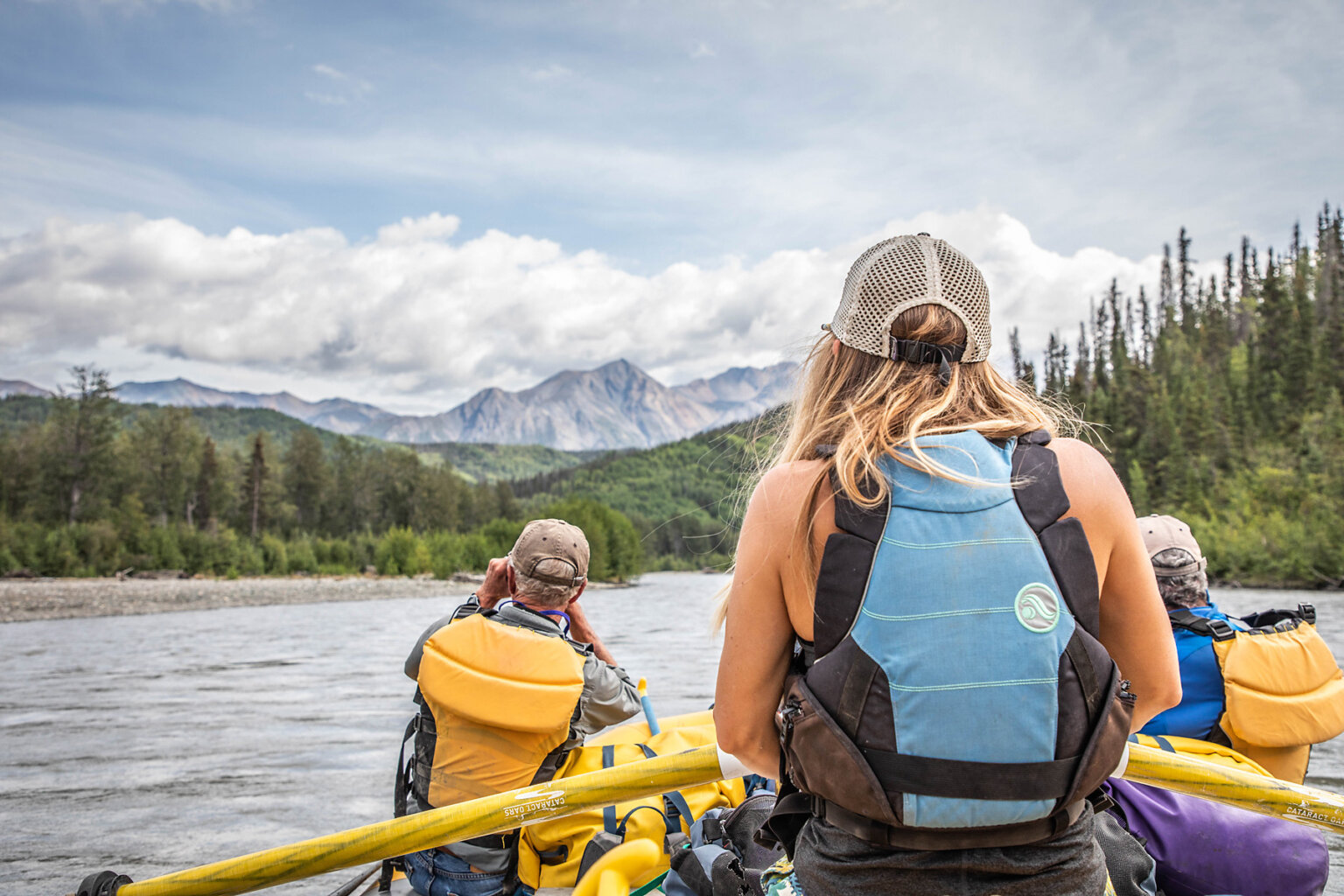 A guide rows while two people in the front of the raft take in rolling green forrested hills and giant mountains downstream on the Tatshenshini River.