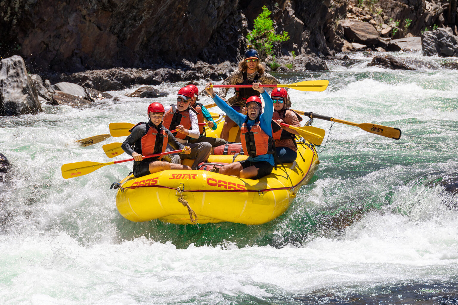 Five guests and a guide on a yellow raft as they're about to enter a rapid while one guests holds his paddle up in the air in excitement.