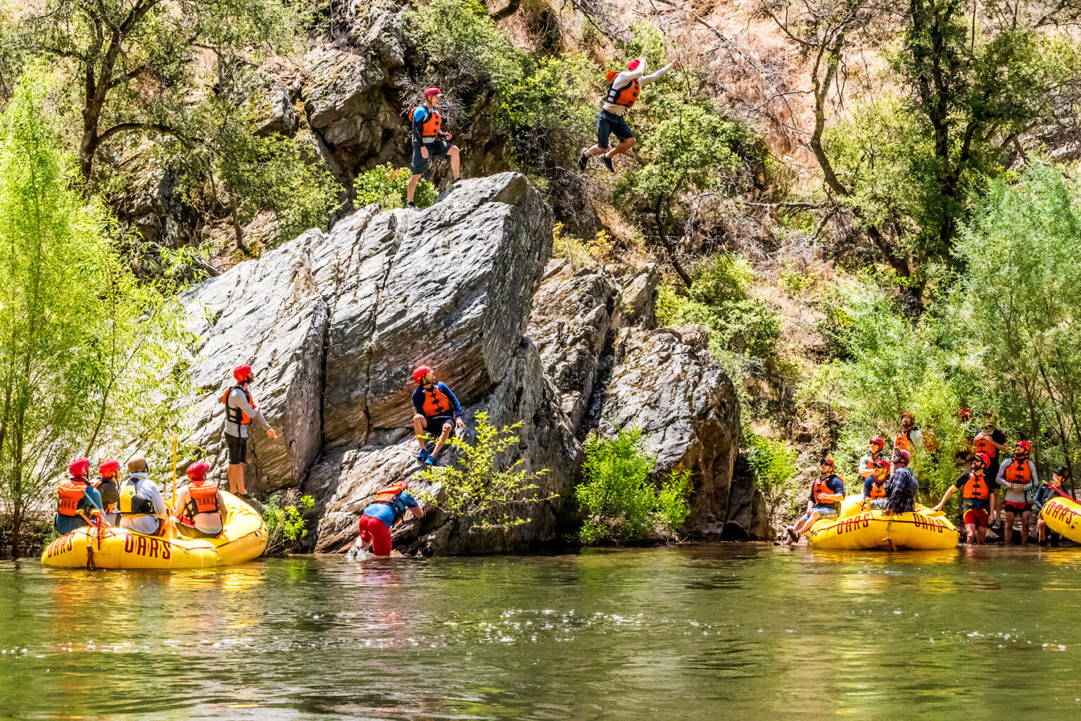 Several guests sitting in yellow rafts watch as other guests jump into the Tuolumne River from a large boulder.