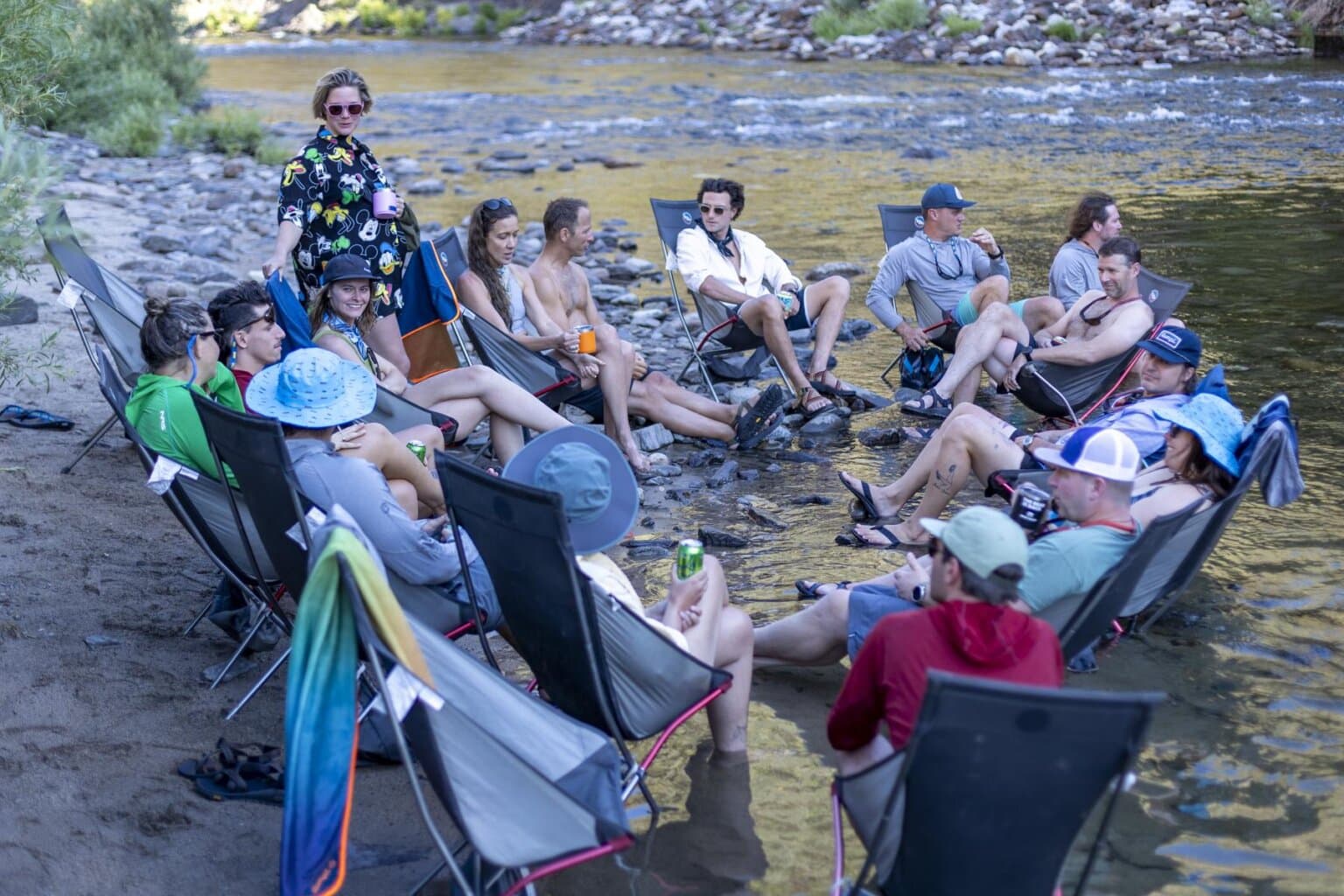 A group of people sit in camp chairs by the Tuolumne River on an OARS rafting trip