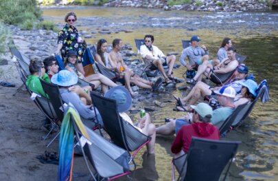 A group of people sit in camp chairs by the Tuolumne River on an OARS rafting trip