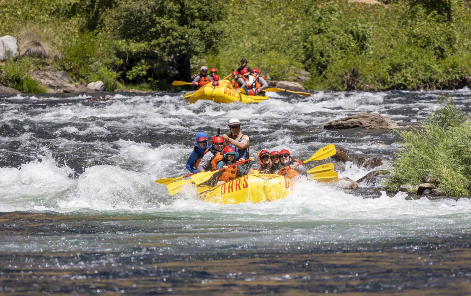 Two yellow rafts navigate through swift river rapids, surrounded by lush greenery under a bright sunny sky.