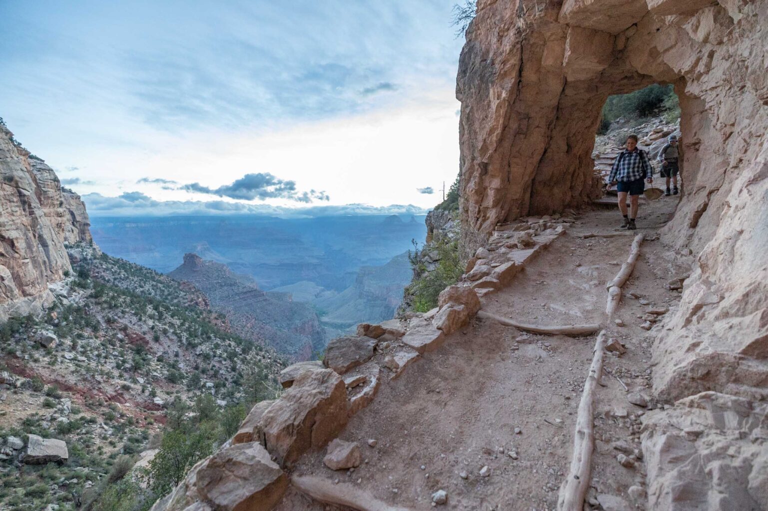 2 people walking through a hole in the rock on a trail next to a cliff.