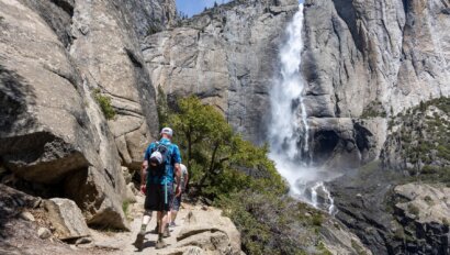 Two hikers in Yosemite National Park on a rocky trail with stunning Yosemite Falls cascading down a towering cliff in the background, surrounded by greenery.