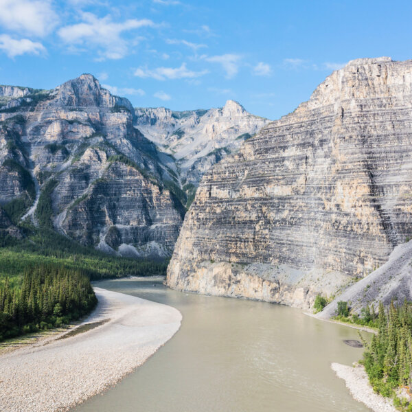 A wide view of forested green hillside, rocky beaches, and giant mountains surounding the Nahanni River in Alaska.